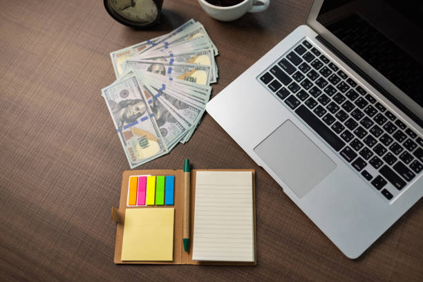 Office workspace featuring a laptop, alarm clock, coffee, colorful stationery, and a pile of hundred-dollar bills.
