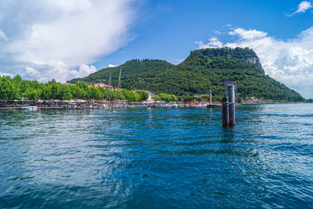 A general view of the harbour of Garda on Lake Garda. The blue skies are reflected in the water. The scene shows the shoreline with various outside cafes and the rocky hills that rise above the town.