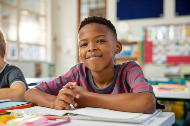 Portrait of african american child sitting at desk at elemetary school Portrait of happy black elementary boy studying in classroom while looking at camera. Close up face of little satisfied black boy writing in notebook using pencil. Proud schoolchild studying in classroom in primary school. young black boys stock pictures, royalty-free photos & images