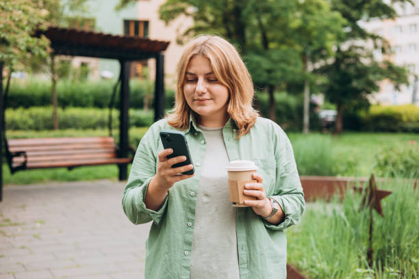 istock Beautiful smiling 30s female with coffee cup using smartphone and standing outdoors. Phone Communication. Happy cheerful young woman walking on city park, Urban lifestyle concept. 2160475559