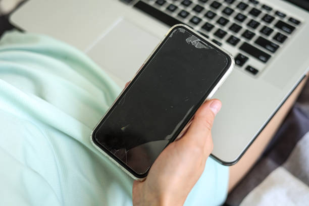 istock Unrecognisable woman is using, texting, messaging smartphone with a broken touch screen, glass with cracks. Black mockup. Laptop on background. Replacing broken glass on a cell phone. 2160478086