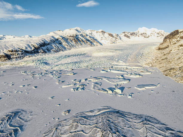 Aerial view of the Skaftafell Glacier in Iceland, showcasing expansive ice fields and rugged mountain ranges under a clear blue sky. The landscape features intricate ice patterns and textures, highlighting the glacier's vast and frozen terrain.