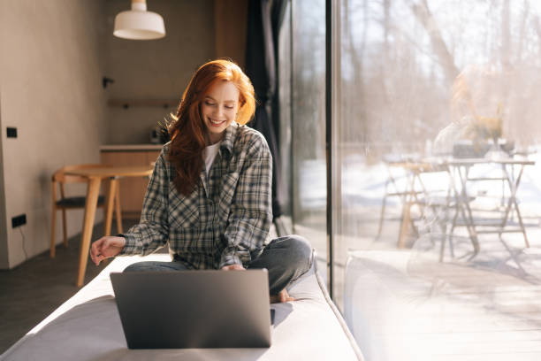 istock Front view of happy freelancer female remote working on laptop computer sitting on wide windowsill with nature view on sunny day. 2160510177
