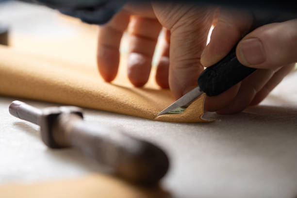 In this photo taken in a shoe workshop, an experienced craftsman over the age of 60 is measuring a piece of leather on the table with his self-made templates and cutting it with a cutting tool. This scene highlights an essential stage in the traditional shoe-making process.