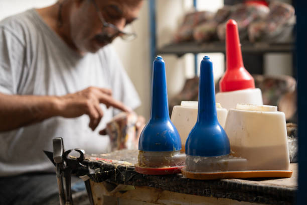 In this photo, a shoe craftsman is seen working behind the bench, applying glue to the sole of a shoe with his finger, planning to attach the sole to the bottom of the shoe. In the background, glue and several shoes with glue applied to their soles can be seen. This scene highlights an essential stage in the shoemaking process and the craftsman’s attention to detail.