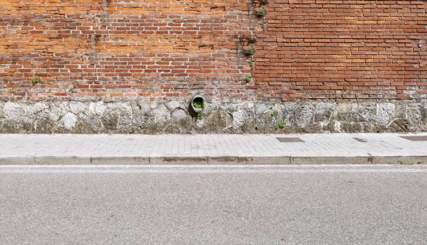 istock Old concrete pipe for water drainage on a retaining brick wall. Tiled sidewalk and urban street in front. 2160555618