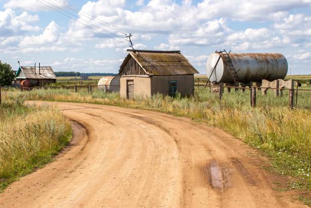 Old farmhouse in the countryside with a dirt road leading up to it, a nearby silo, and rolling fields beyond. A rusty tank is parked in the background next to a utility pole. The sky is partly cloudy.