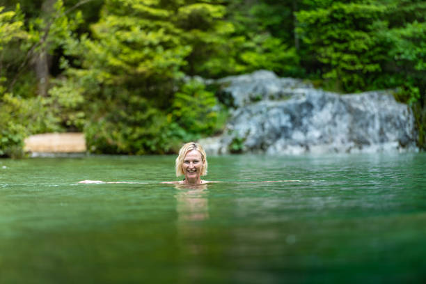 A woman bathes in green cold mountain water. The Wim Hof ​​method, cold therapy, breathing techniques, meditation.Swimming in the pool of the mountain ice river in Kamniška Bistrica. She is in the river, enjoying the outdoors, high concentration, meditation. A green river in an emerald green cove in the forest.