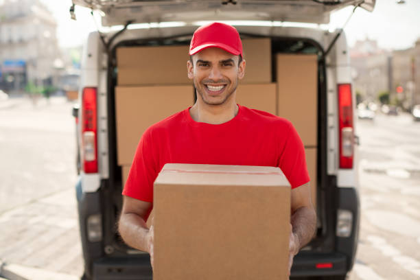 istock Happy young courier man in red uniform standing with parcel box near car outdoors and smiling at camera 2160590342