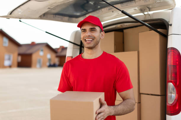 istock Young delivery man in uniform looking and smiling at camera while holding package, standing near van full of cardboard boxes 2160593788