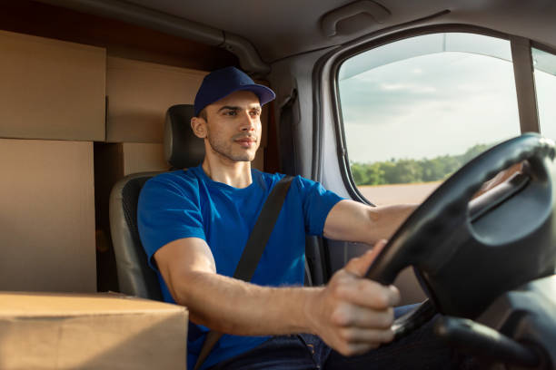 istock Young serious delivery man in uniform sitting inside van full of cardboard boxes, delivering parcels to clients 2160594111