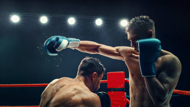 istock Two Male Boxers in an Electrifying Match, One Delivering Powerful Punch Under Bright Lights. Intensity and Raw Energy of the Fight Are Captured, With Sweat Flying Off Their Bodies. Boxing Championship 2160594910