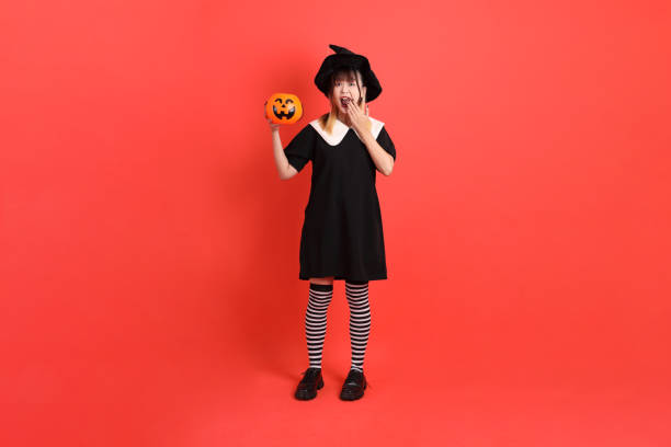 Young asian woman in witch Halloween costume with gesture of Fear and surprise to the hand holding Jack-o-Lantern Pumpkin to her isolated on the orange background. October.
