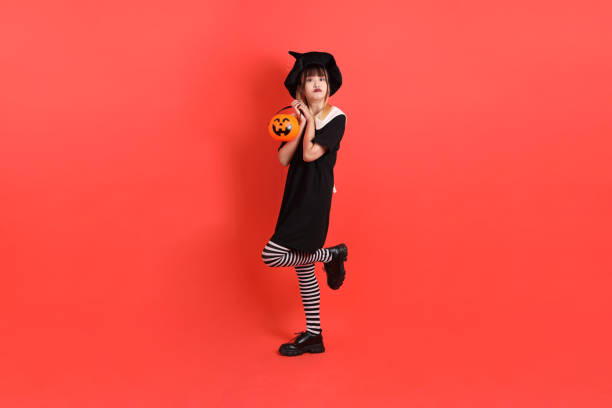 Young asian woman in witch Halloween costume with gesture of smiling and standing while  holding Jack-o-Lantern Pumpkin  isolated on the orange background. October.