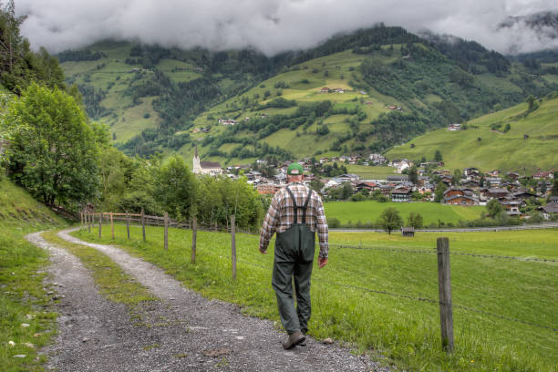 A hiker goes down from the alpine pasture into the valley. In front of him lie the houses of the market town Rauris in Austria. The surrounding mountains are shrouded in thick clouds.