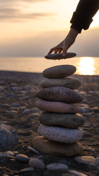 istock Close up woman's hand puts the last stone in the pyramid, copy space. Balanced pyramid of pebbles on the beach against the background of the sea at sunset. Vertical photo 2160601397