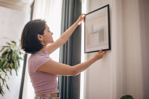 Woman hanging framed abstract art on white wall. A woman in a purple top and jeans is seen hanging a framed abstract artwork on a white wall. The setting appears to be a modern, well-lit living space with plants visible in the background. hanging art on wall stock pictures, royalty-free photos & images