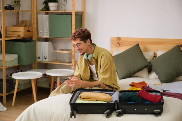 istock Young man sitting on bed packing suitcase using smartphone 2160611214