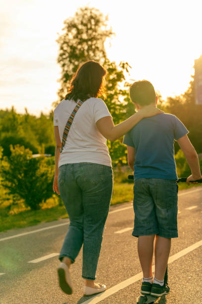 Mom and son walk on a paved path, with the sun shining warmly overhead.  scooter