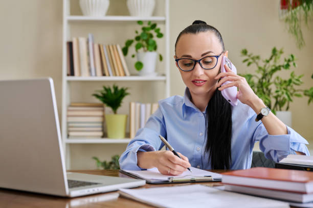 istock Business woman talking on mobile phone sitting at desk with computer 2160631405