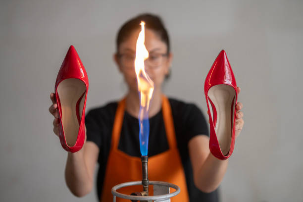 A young woman is seen sitting at a workbench in a shoe workshop. She is wearing an orange apron and holding a shoe in her hand. Various equipment used in shoemaking, including leather, templates, cutting tools, and glue, are on the workbench. This scene highlights the meticulous stages of the shoemaking process.