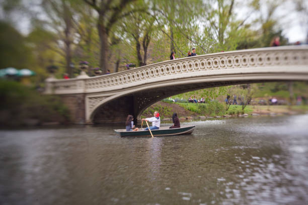New York City, New York, USA - 07.31.2024: Several rowboats glide across the lake under the iconic Bow Bridge in Central Park. Lush green trees and calm water create a peaceful scene in the heart of New York City.