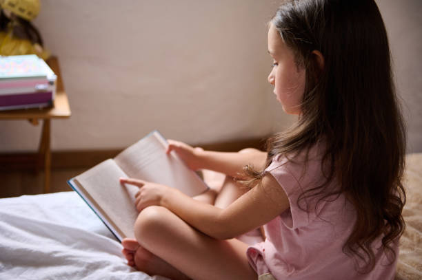 istock View from the shoulders to a cute elementary age student girl reading book, running her finger along the sentences line, enjoying fairy tale in the morning, sitting on the bed in her cozy bedroom 2160634687