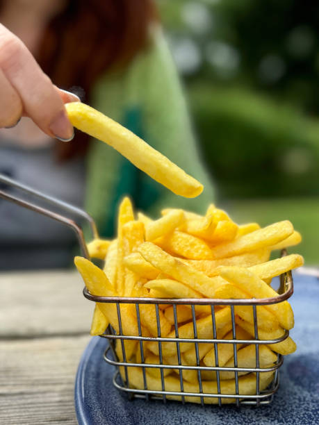 Stock photo showing close-up view of a metal basket of French fry frites on a plate in an al fresco dining setting. imagen de una mujer pelirroja irreconocible tomando papas fritas de la canasta de metal de papas fritas en un plato, sosteniendo alevines entre el dedo índice y el pulgar, mesa de comedor de madera al aire libre, comida de hamburguesa de pollo y papas fri