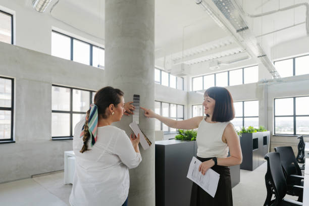 Photo of two woman, coworkers in the same company, choosing a paint shade for a wall in their new office premises,  from a colour palette (colour swatch)