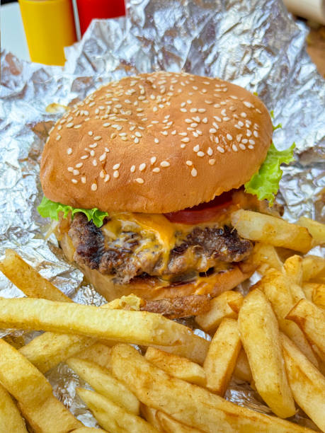 Stock photo showing close-up view of a gourmet cheeseburger in a sesame seed bread bun, with a tasty grilled beef patty, melted cheese slice, tomato slice and lettuce, sitting on aluminium foil. imagen de primer plano de papas fritas y hamburguesa con queso gourmet en papel de aluminio, hamburguesa de carne de res de comida rápida en pan de semillas de sésamo, rebanada de queso derretido, rebanada de tomate, lechuga, mostaza y salsa de tomate, f