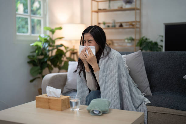istock Woman Sneezing into Tissue While Sitting on Sofa at Home, Surrounded by Plants and Blankets, Highlighting Common Cold and Flu Symptoms 2160693963