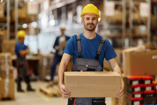 Wide shot of a Caucasian female warehouse worker puling a pallet jack stacked with cardboard boxes.