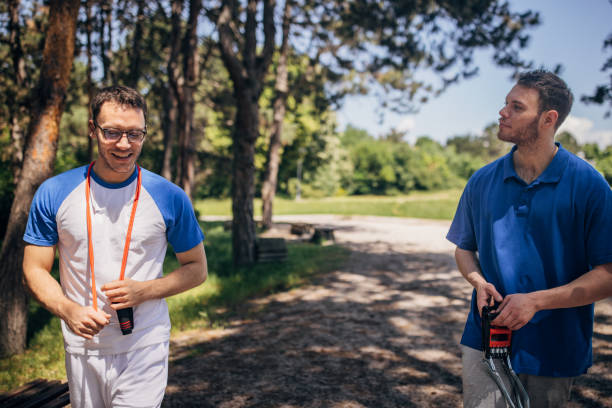 Two friends are walking in a sunlit park. One man is holding a jump rope while the other, wearing a blue polo shirt, walks beside him engaging in casual conversation.