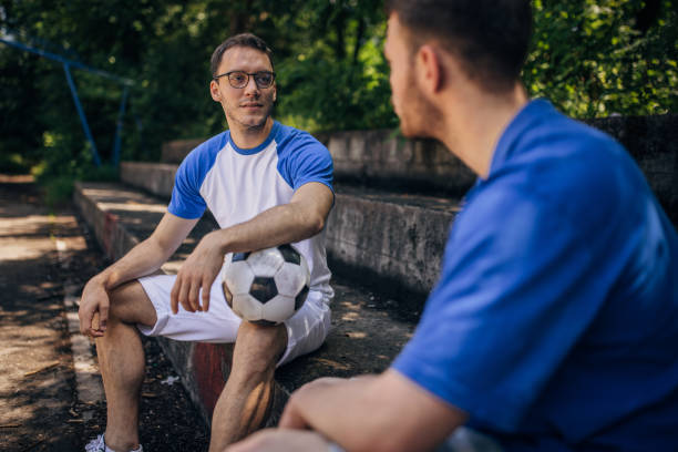 Two young men, one holding a soccer ball, are sitting on bleachers and having a conversation in a park while surrounded by lush greenery.