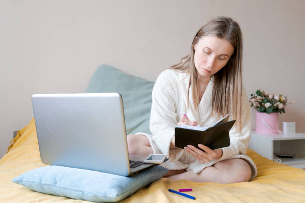 istock A woman is sitting on a bed with a laptop and a notebook 2160713914