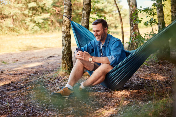 istock Man sitting in hammock using mobile phone in forest 2160714233