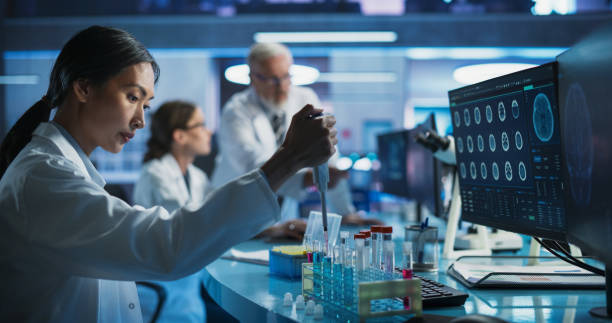 istock Biotechnology Research Laboratory: Female Asian Doctor Working With Test Tubes To Study Effect Of Innovative Alzheimer's Medicine. Focus Shifting To Colleagues Discussing CT Brain Scans On Computer. 2160715202