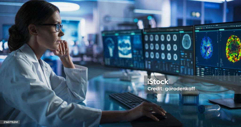 Medical Research Center Laboratory: Caucasian Female Neurologist Using Desktop Computer With MRI And PET Scans Of a Brain. Professional Doctor Looking For Areas of Patient's Brain Damaged By Stroke. Healthcare And Medicine Stock Photo