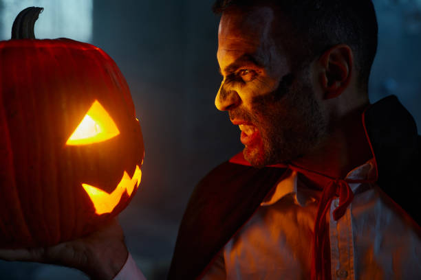 Scary man in count Dracula costume facing Jack O' Lantern on Halloween.