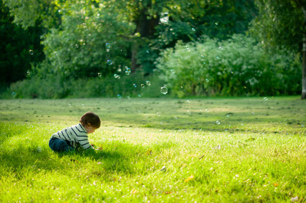 A small child is sitting on the grass playing with soap bubbles outdoors. It's a sunny day