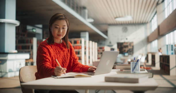 Diligent Asian Female Student Engaged in Academic Research With Laptop and Notebook in Modern Library. Young Woman Focused on Studying, Surrounded by Books and Educational Materials.
