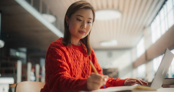 istock Concentrated Asian Female Student Working on a Project With Her Laptop in a Modern Library. Young Woman Writing Notes, Engaged in Academic Research and Study Preparation. 2160740621
