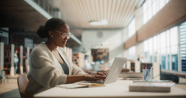istock Dedicated African American Female Student Engaged in Online Learning at a Modern Library. Young Woman Using Laptop for Research, Surrounded by Books and Academic Environment. 2160740658