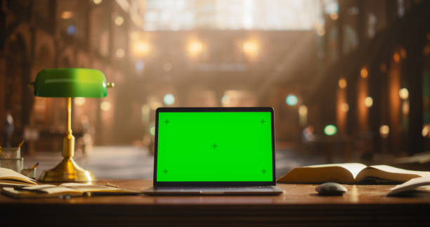 istock Laptop Computer with Green Screen Mock Up Chromakey Display Standing on a Wooden Table in an Academic Traditional Library. Office Desk With Books, Notebooks, Lamp and Wireless Mouse 2160741583