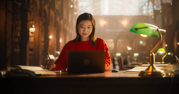 istock Dedicated Asian Female Student Engaged in E-Learning with Laptop in Classical Library. Young Woman Concentrating on Studies, Surrounded by Books, in a Cozy, Well-Lit Study Environment 2160741813