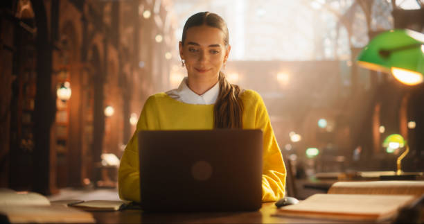 Engaged Caucasian Female Student Using Laptop for Online Learning in a Cozy Library. Young Woman Concentrated on E-Learning, Surrounded by Books and Warm Ambiance