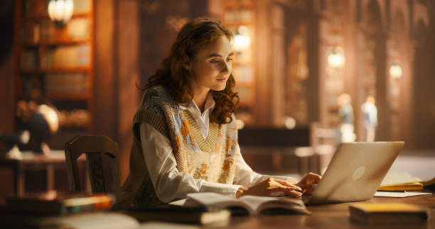 istock Serene Young Woman Engaged in Studying with Books and Laptop in a Classical Library Setting, Illuminated by Warm Ambient Light. Caucasian Female Focused on Academic Research and E-Learning. 2160742157