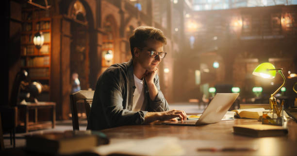 Focused Young Man Typing on Laptop in a Majestic Library, Illuminated by Soft, Warm Light, Symbolizing Knowledge, Creativity, and the Pursuit of Literary Excellence.