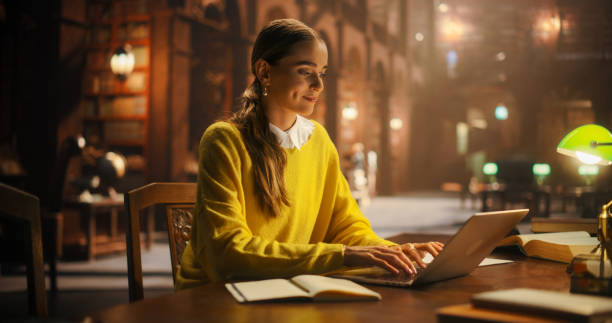 istock Focused Young Woman Studying in a Classic Library, Using Laptop for Online Education. Engaged in E-Learning, She Prepares for University Exams by Reading and Taking Notes. 2160742826