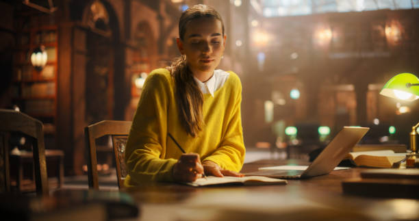 istock Focused Young Woman Studying In a Vintage Library, Surrounded by Books, Using a Laptop For Research and Writing in Notebook. Calm Academic Setting Highlights a Dedication to Learning and Education 2160742829
